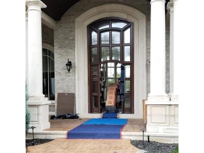 A professional mover is shown carefully navigating a stack of large boxes through the grand, arched wooden entryway of a luxury estate. The entrance features massive white columns, a tall vaulted ceiling, and protective blue floor runners leading from the