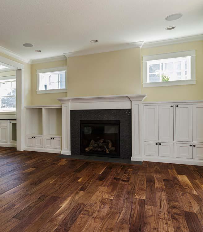 The image shows a beautifully refinished hardwood floor in a modern living room. The wood planks have a rich, warm tone with a glossy finish that highlights the natural grain and texture. The room is well-lit, with sunlight streaming in from large windows