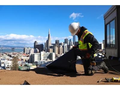 A construction worker in a white hard hat and a high-visibility yellow safety vest kneels on a flat rooftop, focused on applying a black roofing membrane. The worker is using a specialized tool to secure the material onto a tan, textured surface. The scen