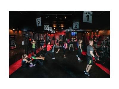 The image shows a gym with multiple people engaging in boxing and fitness exercises under numbered "Round" signs. The dimly lit space has a red and black color scheme, creating an intense workout atmosphere.