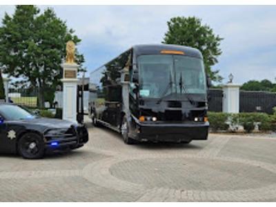 A sleek black tour bus arrives at an elegant gated entrance, accompanied by a police vehicle, adding a sense of security and importance to the scene.