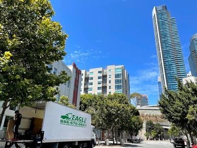 A moving truck is parked on a city street with high-rise buildings and trees in the background. A worker is inside the truck, organizing or unloading items. The bright blue sky and urban setting highlight the moving process in progress.