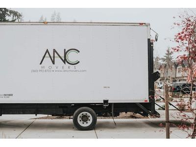 A white moving truck with the "ANC Movers" logo is parked on a driveway. The branding on the side includes the company's name, phone number, and website. The background features a suburban neighborhood with houses, trees, and parked cars, creating a pr