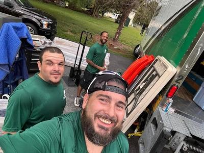 Three movers wearing matching green shirts are taking a selfie while unloading or loading a moving truck. They appear cheerful and friendly, with one man in the foreground smiling broadly. The background includes a truck with a green and white design, mov