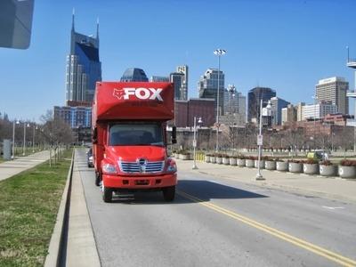 A red moving truck is parked on a road with a city skyline in the background, featuring modern high-rise buildings under a clear blue sky. The bold design on the truck highlights a professional moving service.