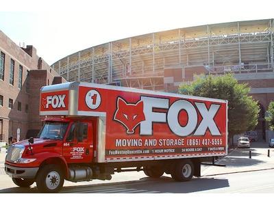 A bright red moving truck is parked near a large stadium, standing out against the urban backdrop. The bold branding and contact details emphasize reliability and service availability.