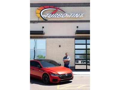 An image of a man standing with his arms crossed next to a sleek red Volkswagen with dark window tinting, showcasing the work of Turbo Tint, a premier provider of car window tinting services. The scene is captured outside the brick storefront, which promi