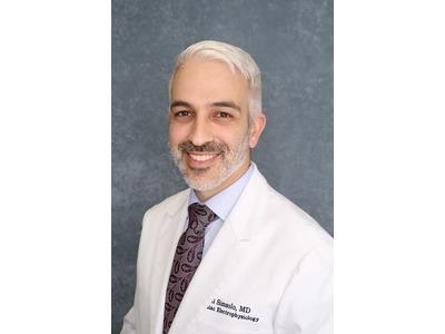 An image of a professional medical headshot featuring Eli Simsolo, MD, a highly regarded electrophysiologist LA (Los Angeles) specialist. Dr. Simsolo is pictured smiling in a white lab coat embroidered with his name and specialty over a light blue collare