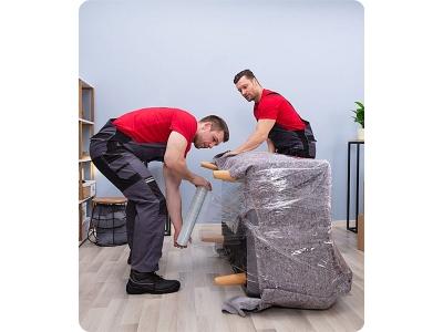 Two professional movers from Rest & Relaxation Moving/Delivery LLC are carefully wrapping a padded chair in protective plastic film. Dressed in matching red shirts and gray work pants, they demonstrate precision and teamwork in a clean indoor setting, emp