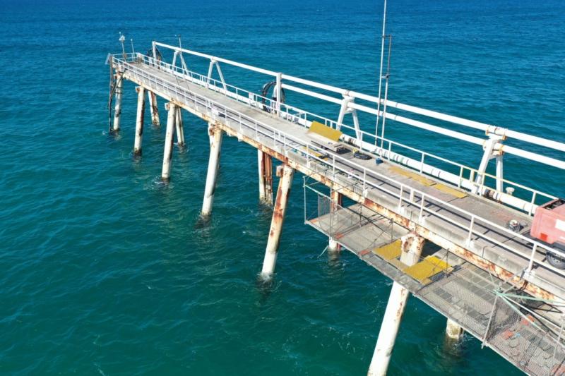 Aerial view of a long steel jetty extending over deep turquoise water; rust-streaked support piles and a suspended work platform illustrate the challenging access environment.