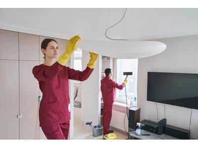 Two Sparkle Janitorials cleaners in maroon uniforms clean a modern living space, one wiping a ceiling light fixture while the other uses a squeegee on a window.