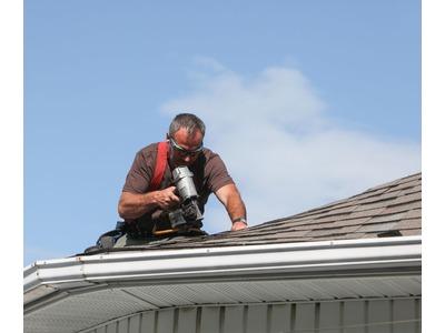 An image of a focused roofing professional from Premier Home Solutions working on a shingled roof, using a nail gun under a clear blue sky.