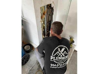 Image of a plumber from Plumbing Around the Clock at work, seen from behind, focusing on an exposed section of wall plumbing, wearing a company shirt with contact information in Fort Lauderdale, FL.