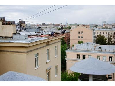 View of two workers on a rooftop in an urban environment. They are engaged in maintenance work, surrounded by roofing materials and tools. The architecture of the surrounding buildings is classic, with a mixture of residential and possibly commercial stru