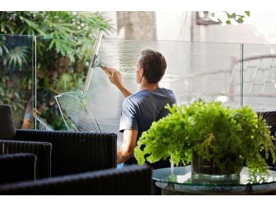 The image shows a man cleaning a large glass window in a well-lit indoor setting that appears to be a lobby or a lounge area. He is using a squeegee to wipe the window. The surrounding area features lush greenery, including a potted plant on a coffee tabl