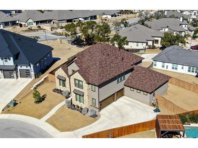 The image is an aerial view of a large, two-story house in a suburban neighborhood. The house features a combination of stone and stucco exterior walls with a complex, multi-gabled roof covered in brown shingles. The property includes a neatly landscaped 