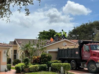 The image captures a roofing team at work on the roof of a one-story suburban house. Three workers, wearing bright safety vests, are either inspecting or repairing the roof. Beside the house, a large red dump truck is parked, likely there to collect debri