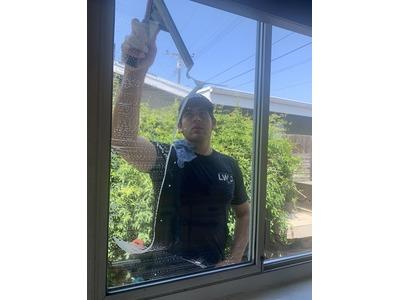 The image shows a man cleaning a window from outside, wearing a dark shirt and using a squeegee. The background reveals a residential area with greenery, emphasizing his focus and professionalism.