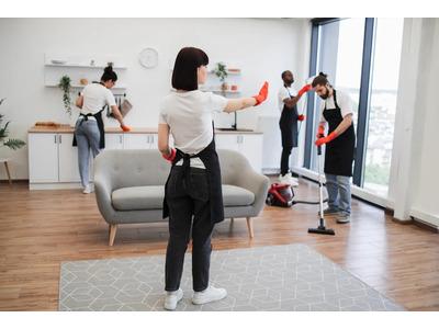 The image shows a team of four professional cleaners working in a modern, well-lit living room. One woman in the foreground is organizing the cleaning process, directing her team, while her colleagues are engaged in different tasks: vacuuming the carpet, 