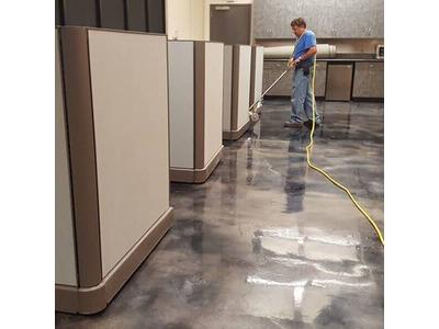 A janitor from Clear Choice Janitorial meticulously cleans a large commercial kitchen floor using a floor buffer connected to a yellow power cord. The reflective sheen on the polished dark floor highlights the effectiveness of their cleaning services, und