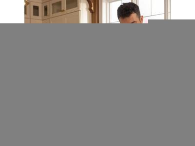 The image shows a man in a dark blue polo shirt and gloves, carefully examining a food sample on a kitchen counter. The kitchen is well-equipped, featuring white cabinetry and a variety of food items and drinks spread out, including orange juice and fresh