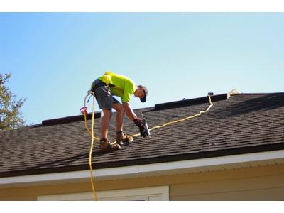 A construction worker in a bright yellow safety vest and helmet is securing shingles on a steep, dark-shaded roof of a house. The worker is using a nail gun connected to a yellow cord that trails across the roof, emphasizing safety and efficiency in the s
