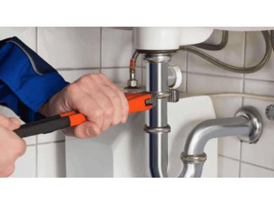 A close-up of a plumber's hands using an orange wrench to adjust a pipe under a kitchen sink. The plumbing includes a shiny silver pipe connected to the sink's drain and other fixtures. The plumber is wearing a blue uniform, focusing on ensuring a tight