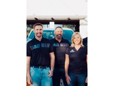 A group photo of three smiling employees from Spartan Home Services, dressed in matching black polo shirts with company logos. The two men and one woman are standing in front of a blue company van, suggesting they are part of a professional team, possibly