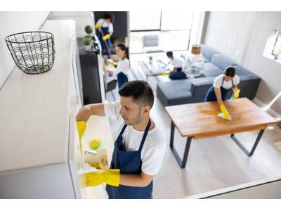 A team of four professional cleaners wearing white shirts, blue aprons, and yellow gloves are cleaning a modern living space. One person is cleaning the inside of a cabinet, another is wiping down a dining table, and two others are working in the backgrou