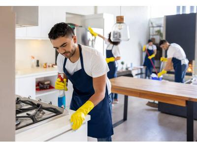 A team of four professional cleaners, wearing white shirts, blue aprons, and yellow gloves, is cleaning a modern kitchen and dining area. One person is cleaning the stovetop, another is wiping down a table, while the other two are working in the backgroun