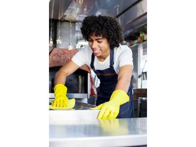 A smiling worker with curly hair, wearing a white shirt, blue apron, and yellow gloves, is cleaning a stainless steel surface in a commercial kitchen. The scene suggests professional kitchen cleaning in a well-maintained and organized environment.