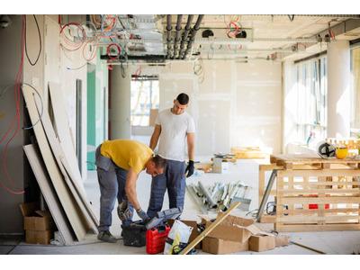 Two construction workers are inside a building under renovation. One worker in a yellow shirt is bending over a toolbox, while the other in a white shirt stands nearby. The room is filled with construction materials, tools, and wiring hanging from the cei