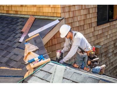 A construction worker in a white hard hat and grey shirt is installing shingles on a roof, using a nail gun. Various tools and materials are visible around him, with cedar shake siding on the adjacent walls.