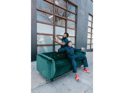 Two movers in dark work uniforms are handling a green velvet sofa outside a building with large windows. One mover is standing behind the sofa, holding a strap, while the other is sitting on the sofa, spreading his arms and smiling. The scene is set on a