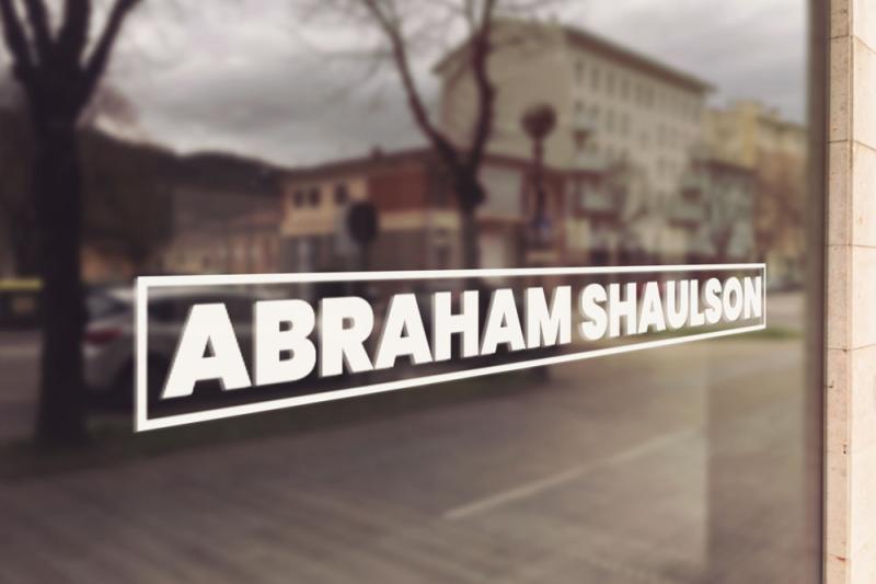 The image shows a logo on a window, featuring the name "ABRAHAM SHAULSON" in bold, white capital letters. The text is enclosed within a rectangular border. The background is slightly blurred, showing an outdoor scene with trees and buildings.
