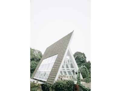 A-frame house with a steep, triangular roof covered in dark shingles, featuring large glass windows and surrounded by lush greenery.