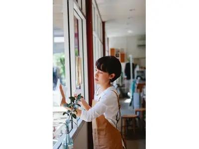 A young woman wearing an apron, standing near a window and looking outside thoughtfully, capturing a moment of reflection and serenity in a cozy, well-lit space.