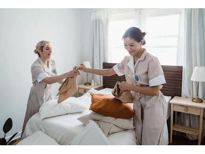 Two housekeeping staff in beige uniforms making a bed with fresh linens and arranging pillows in a bright, tidy room, demonstrating their commitment to maintaining cleanliness and comfort in guest accommodations.