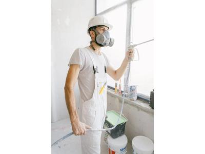 A renovation worker wearing a white hard hat, protective goggles, and a respirator mask, holding a spray gun while preparing to paint a room, emphasizing safety and precision in renovation work.