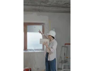 A construction professional wearing a white hard hat and holding a tablet, pointing at the ceiling of a room under renovation, illustrating the planning and inspection process involved in home improvement projects.