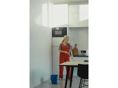 A woman wearing red overalls is mopping the floor in a modern kitchen. A blue bucket is placed nearby, and the kitchen features minimalistic decor with white walls, a refrigerator, and a table with chairs.