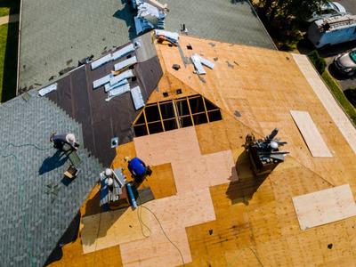 An aerial view of a roofing project in progress, showing several workers installing new shingles on a house roof. Part of the roof is covered with wooden panels, while other sections have black underlayment and new shingles. Tools and materials are scatte