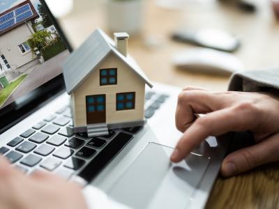 A person using a laptop with a miniature house model placed on the keyboard, symbolizing online real estate searches or transactions.