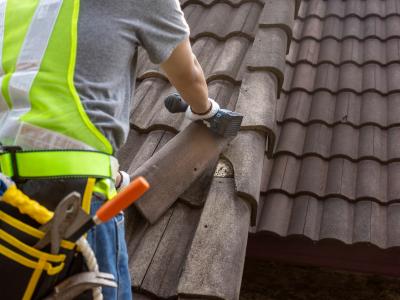 A roofer wearing a safety vest and tool belt is installing clay tiles on a roof. The worker is using gloves and carefully placing the tiles, ensuring they fit properly. The scene highlights the meticulous work involved in roofing installation.