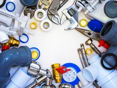 An assortment of plumbing tools and fittings arranged in a circle on a white surface, including wrenches, pipes, connectors, and tape.
