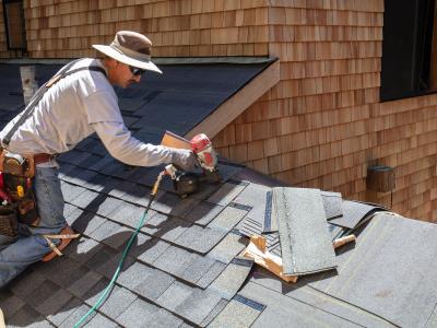 A roofer wearing a hat and sunglasses is installing asphalt shingles on a roof using a roofing nailer. He has a tool belt and various materials around him, working on a house with wood shingle siding. The scene shows the detailed process of roofing instal