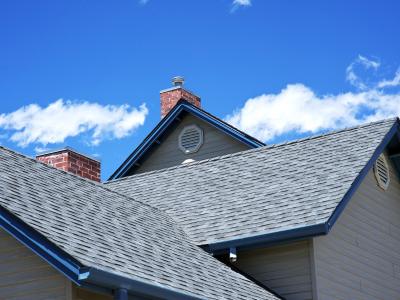 A view of a house with a newly installed gray shingle roof. The roof features multiple gables and two brick chimneys. The sky is clear with a few white clouds, emphasizing the fresh and well-maintained appearance of the roofing work.