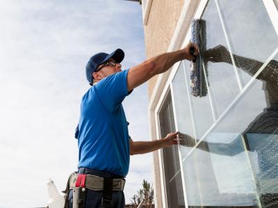 The image shows a man cleaning an outdoor window. He is wearing a blue shirt and cap, and using a squeegee to clean the glass. The background shows part of the building's exterior and a clear sky. The man appears focused on his task, and the reflection i