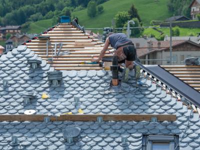 A roofer is working on a slate tile roof in a residential area. The worker is kneeling and using tools to install the tiles, with various roofing materials and tools scattered around. The background shows houses and green hills, highlighting the picturesq