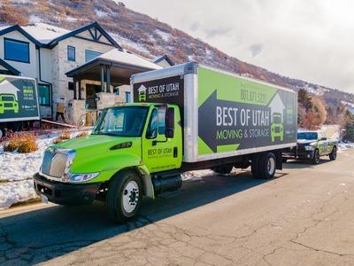 The image shows a green "Best of Utah Moving & Storage" truck parked in a snowy neighborhood with mountains in the background. The truck's signage includes their contact number, 801.871.5221, and the company's name prominently displayed. There's also